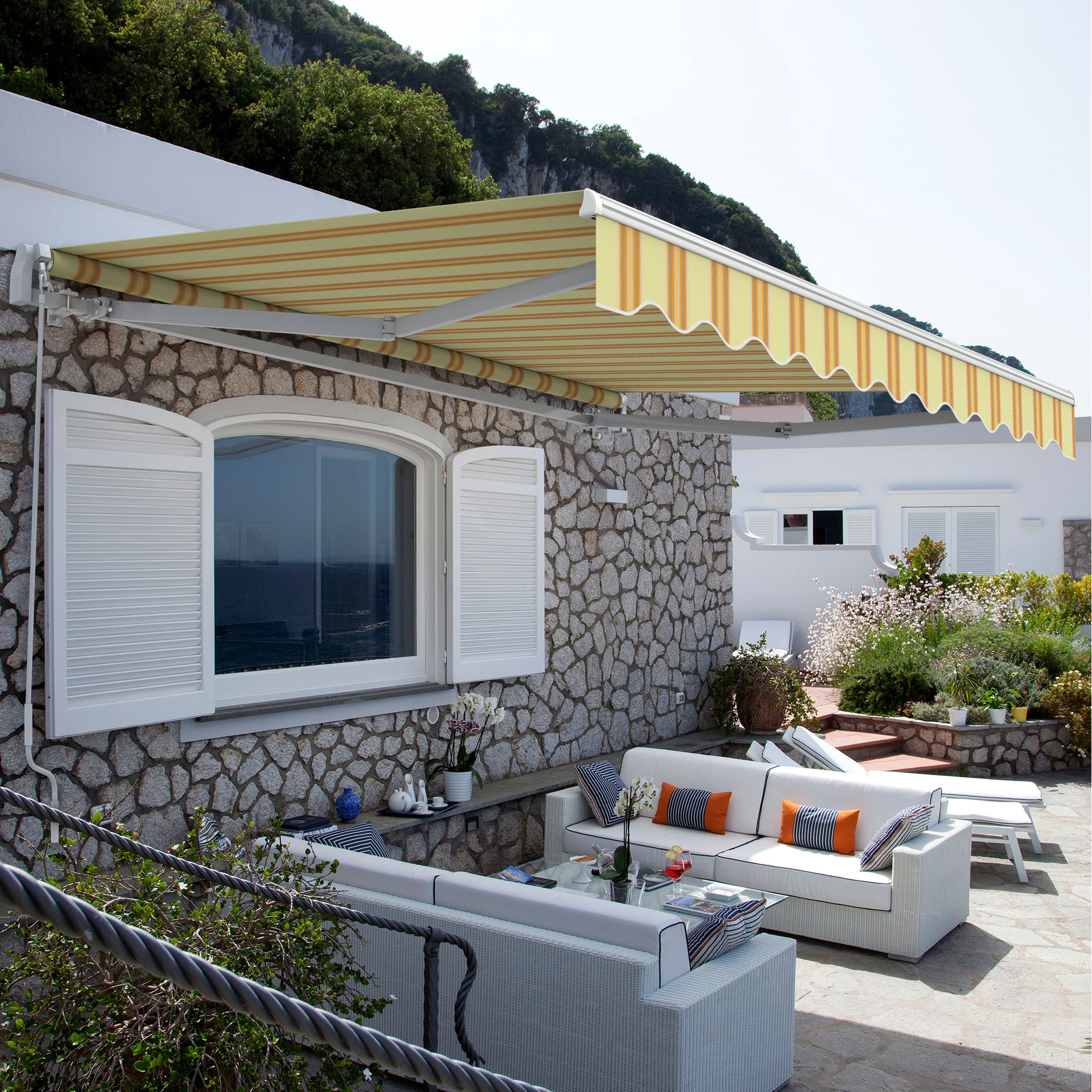 Terrasse mit Markise, Sitzgruppe und Pflanzen vor einem Haus mit Steinmauer und Fensterläden