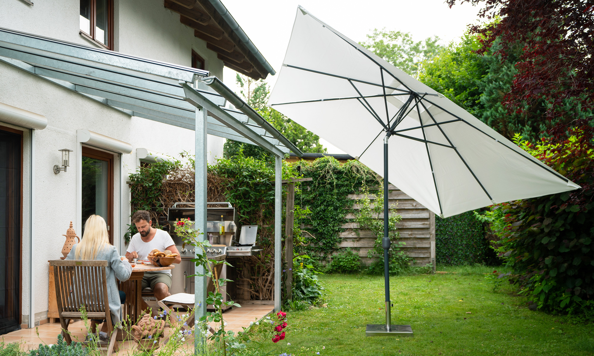 slider_image_13d838ef936b4fc2a64ebb9cd4aa4fca Sonnenschirm mit Kurbelgehäuse im Garten neben Terrasse und Haus, Personen am Tisch sitzend