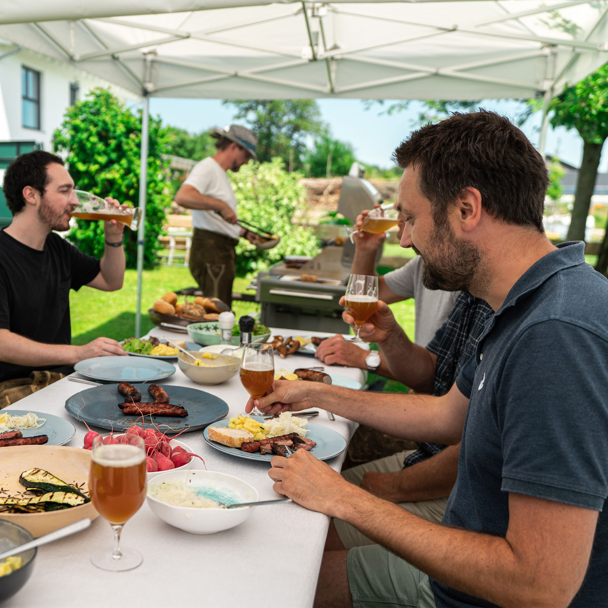 slider_image_a91e52dc3187425ba6100b6e12d19507 Personen sitzen an einem Tisch im Freien und essen, während jemand am Grill steht