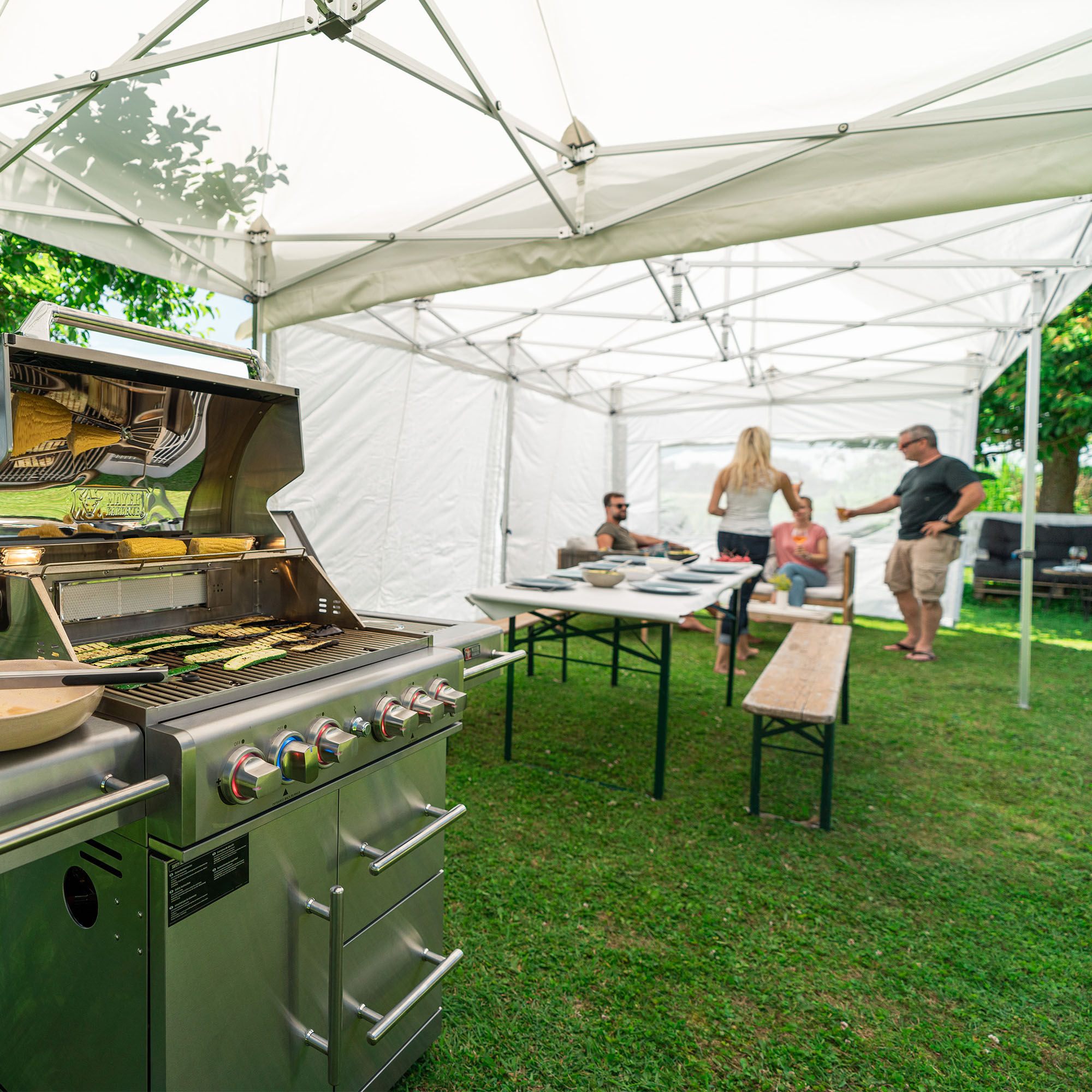 slider_image_69eaaa5aa2c24322ae13a35e4854b83e Grill unter Pavillon mit Tisch und Bänken, Personen im Hintergrund, Gartenumgebung