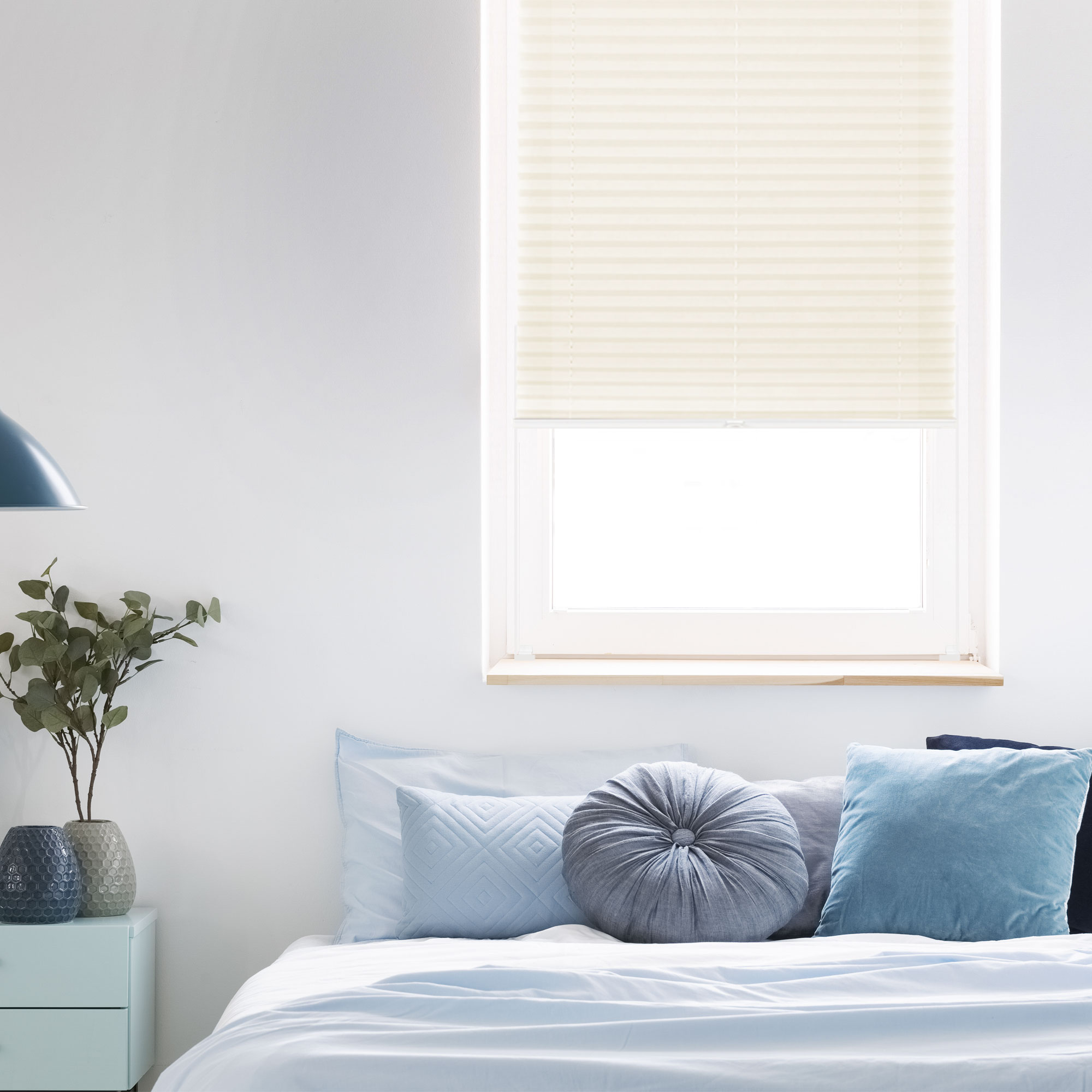 Ein Schlafzimmer mit Bett, Kissen, Nachttisch, Lampe und Fenster mit Plissee-Rollo
