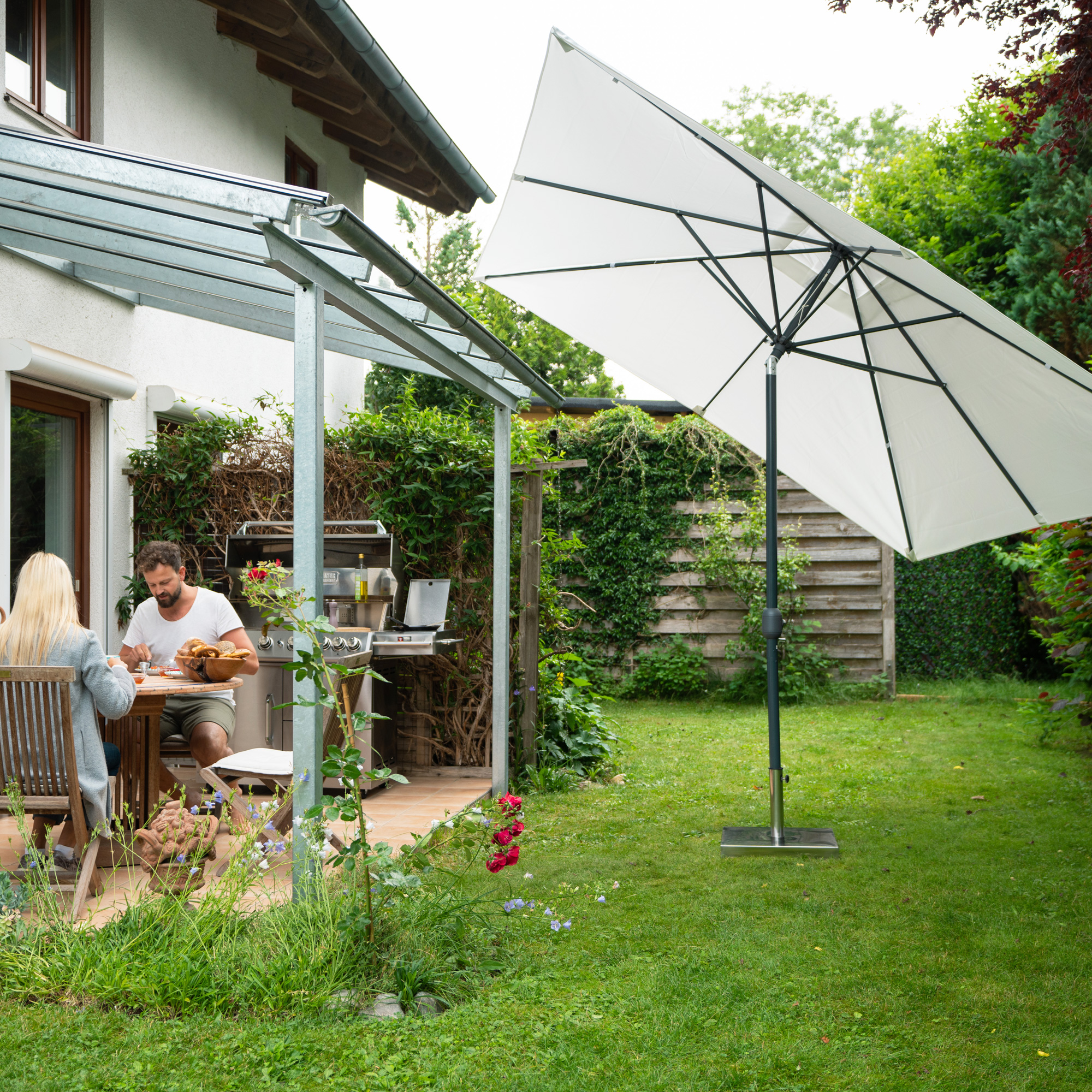 Sonnenschirm mit Kurbelgehäuse im Garten neben Terrasse und Haus, Personen am Tisch sitzend